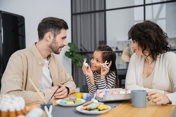 Cheerful girl holding Easter rabbits near parents and food at home.