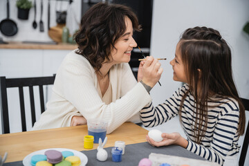 Side view of cheerful mother and child holding paintbrushes near Easter eggs and macaroons at home.