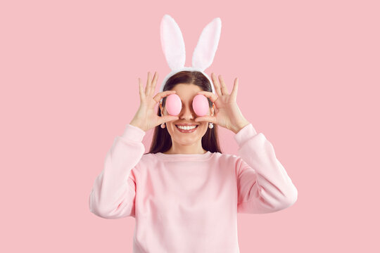Funny joyful brunette young woman in pink sweatshirt with bunny ears on hair band holding easter eggs on eyes on pink background. Happy girl celebrating easter in rabbit costume with pink earrings.