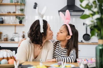Side view of mom and daughter in headbands pouting lips near Easter food in kitchen.