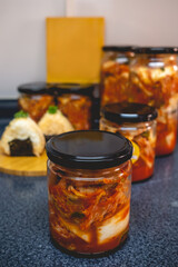 Spicy traditional kimchi in glass jar with kimchi onigiris and other jars in the background (kimchi: mix of pickled and fermented vegetables, garlic, chili peppers, salt) 