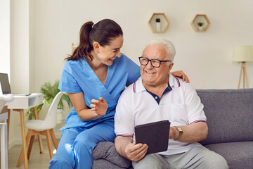 Young friendly female caregiver and senior man using digital tablet together in nursing home. Woman in medical uniform and elderly man are sitting on sofa and discussing modern technology.