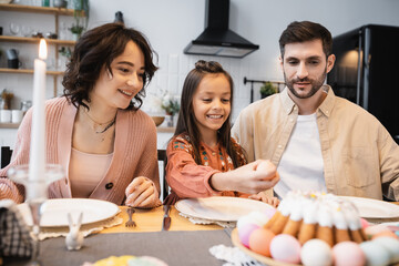 Smiling parents looking at child holding Easter egg during festive dinner at home.