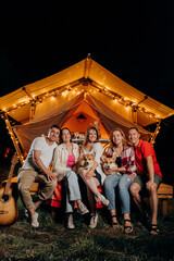 Happy group of friends with Welsh Corgi Pembroke dogs relaxing in glamping on summer evening near cozy bonfire. Luxury camping tent for outdoor recreation and recreation. Lifestyle concept
