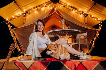 Fototapeta na wymiar Happy young woman with her Welsh Corgi Pembroke dog relaxing in glamping on summer evening near cozy bonfire. Luxury camping tent for outdoor recreation and recreation. Lifestyle concept