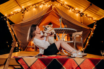 Fototapeta na wymiar Happy young woman with her Welsh Corgi Pembroke dog relaxing in glamping on summer evening near cozy bonfire. Luxury camping tent for outdoor recreation and recreation. Lifestyle concept