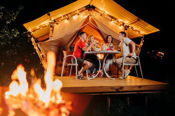 Happy group of friends relaxing in glamping and drinking wine on summer evening near cozy bonfire. Luxury camping tent for outdoor recreation and recreation. Lifestyle concept