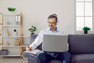 Mature man in headset with laptop working in his home office making notes sitting on couch. Serious focused businessman in headphones on online work meeting. Remote, distant work, job concept.
