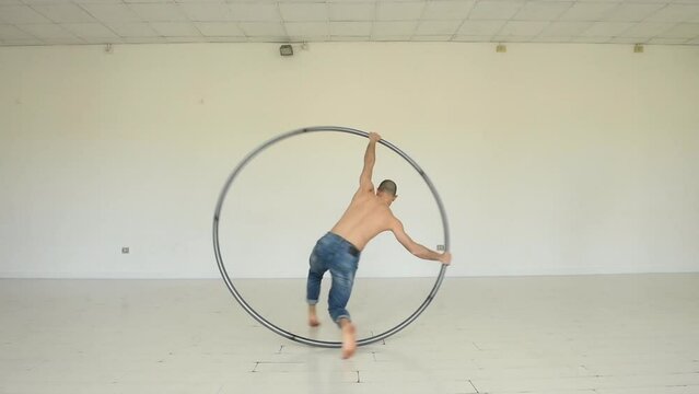 Real time full body of flexible male artist moving around steel cyr wheel then standing on it and hanging while performing different acrobatic tricks in studio 