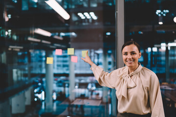 Smiling woman pointing at sticker on glass wall while working on strategy