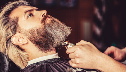 Hands of a hairdresser with a beard clipper, closeup. Bearded man in barbershop. Man visiting hairstylist in barbershop - obrazy, fototapety, plakaty