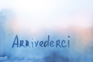 lettering Arrivederci in Italian language is good bye in english on foggy glass on blue sunset window