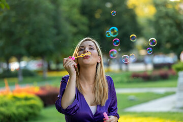 Beautiful Woman with Long Hair and Violet Dress is Walking in Public Park and Holding and Blowing a Bubbles.