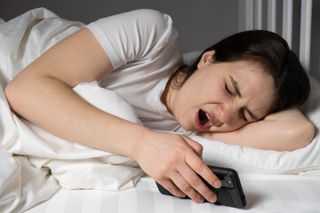 30-year-old woman yawns lying in bed with a smartphone, using a gadget before going to bed
