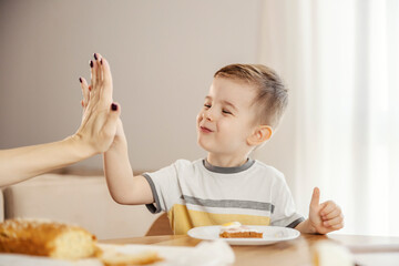 A little boy is sitting with his mom at breakfast table, chewing food and giving high five.
