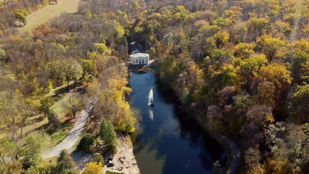 Beautiful panoramic autumn landscape park, many trees with yellow leaves, lake with fountain in center, architecture, big stones, paths walkways, people walking on sunny autumn day. Aerial drone view