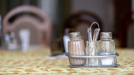Pepper shakers and salt shakers, a napkin stand are on the table in a cafe. The table in the restaurant is covered with a tablecloth. White paper napkins on a stand and a glass container with sugar.