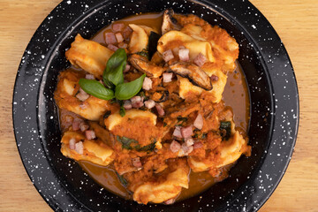 Pasta. Top view of traditional stuffed angelottis with a tomato sauce with mushrooms, bacon and basil in a black dish on the wooden table.