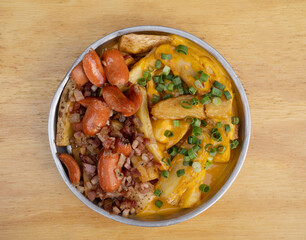 French fries. Top view of fried potatoes with sliced green onion, smoked sausages, crispy bacon and cheddar cheese sauce, in a metal dish on the wooden table.