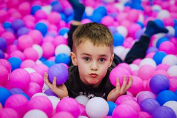 Obraz na płótnie Canvas A beautiful, happy, small, smiling boy, a preschool child lies in a variety of multi-colored, colored plastic balls on the playground. Photography, portrait, childhood concept.