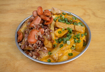 French fries. Closeup view of fried potatoes with sliced green onion, smoked sausages, crispy bacon and cheddar cheese sauce, in a metal dish on the wooden table.