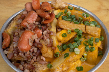 French fries. Closeup view of fried potatoes with sliced green onion, smoked sausages, crispy bacon and cheddar cheese sauce, in a metal dish on the wooden table. 