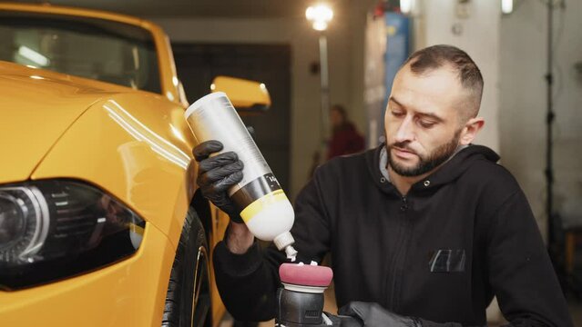 Man auto service worker, wearing black clothes, putting special polish wax or cream on the orbital polisher to polish the luxury yellow car for removing scratches and shine. Putting special polish wax