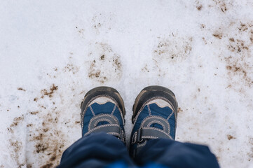 A child, a boy in boots, shoes, stands on the ground with snow in winter outdoors. Close-up photography, lifestyle, top view.