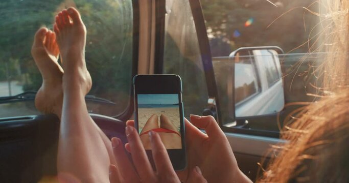 Relaxed woman enjoying road trip getaway in the car looking at photos on her phone on a fun, relaxing and peaceful sunset drive in summer. Barefoot female with her feet up on a holiday break in Italy