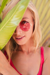 Charming long-haired woman in pink sunglasses in park. Outdoor photo of gorgeous young lady in bright attire touching big green leaf.