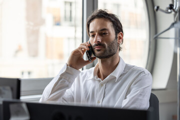 portrait d'un jeune homme souriant, employé de bureau assis devant son ordinateur au bureau. Il téléphone avec son smartphone