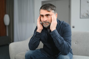 Sad stressed troubled mature middle-aged man sitting on the couch, missing his friends, feeling bad unwell unhealthy, suffering from depression at home