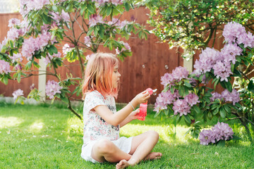 Cute little girl with toned red hair blowing soap bubbles while sitting on a green lawn in the garden. Having fun during school holidays at home. Offline activity for kids. Happy childhood concept