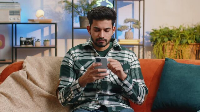 Indian man sitting on sofa uses mobile phone smiles at home living room apartment. Hindu young guy texting share messages content on smartphone social media applications online, watching relax movie