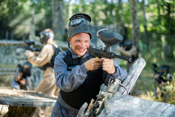 Fototapeta premium Excited caucasian man paintball player standing at cover, aiming with paintball gun and shooting.