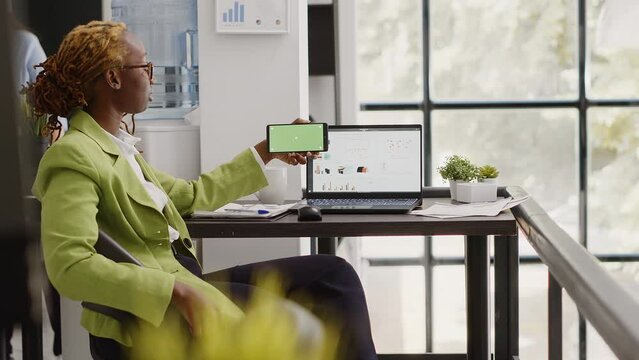 African american worker using horizontal greenscreen on mobile phone, looking at isolated copyspace display in business office. Young employee checking blank chroma key template on smartphone.