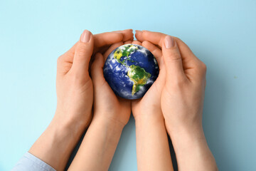 Hands of mother and child holding small planet Earth on light blue background