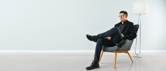 Fashionable young man sitting in armchair near light wall. Banner for design
