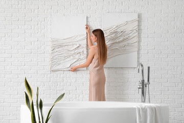 Young woman hanging 3D picture on white brick wall in bathroom