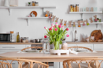 Vase with tulips, Easter eggs, cake and jug of water on dining table in kitchen