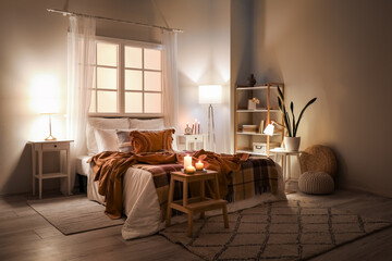 Interior of bedroom with brown checkered blankets on bed, burning candles and glowing lamps late in...