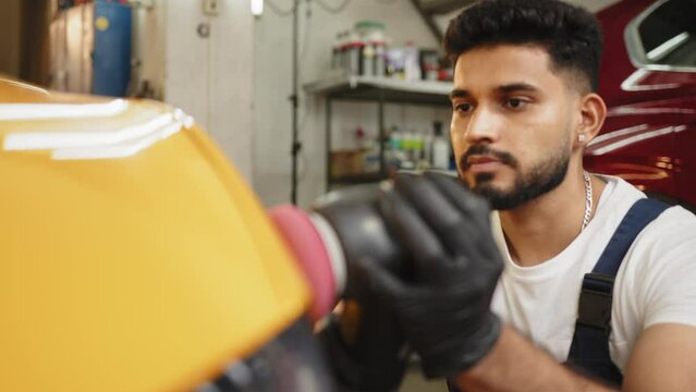 Young handsome bearded man, worker of auto detailing service, holds orbital polisher in the hand and polishes a body and hood of yellow modern car. Car detailing and polishing concept.