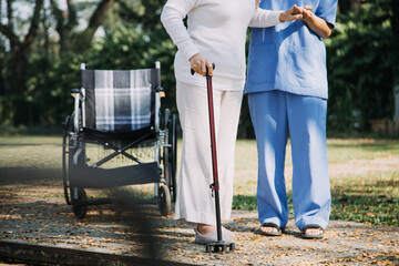 Asian young caregiver nurse support senior older male walking outdoors. Specialist girl doctor help and take care of elderly mature man patient doing physical therapy in public park at nursing home.