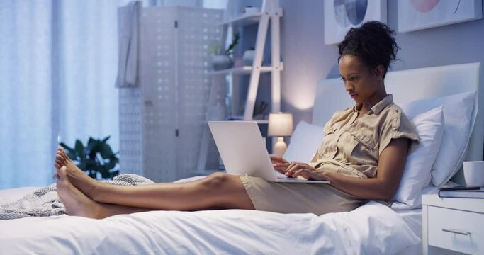 Woman night work from home, laptop in bedroom social media communication, young black girl writing online lifestyle blog and typing email. Internet technology wifi in apartment, working job in house