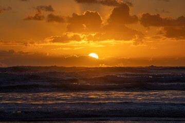 A dramatic orange sunset peeking out from behind a layer of clouds, casting a warm glow over the restless waves of the ocean.