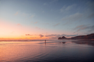 A young woman dances at Bethells beach at sunset, surrounded by vibrant pink, violet, and orange tones.