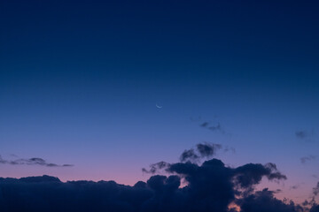 The tranquil beauty of a small crescent moon suspended in a sea of blue and purple hues, peeking out from above the rolling clouds.