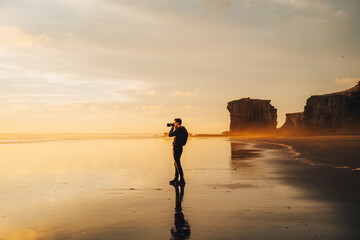 A lone photographer silhouetted against a blazing golden sunset, intently capturing the breathtaking scenery through his lens, at a stunning and secluded beach location.