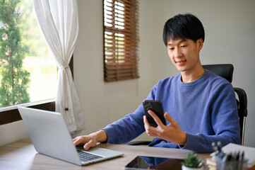 Fototapeta premium Handsome young Asian male office worker using his smartphone at his office desk.