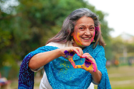 Indian senior woman playing colors and making heart shape with hand